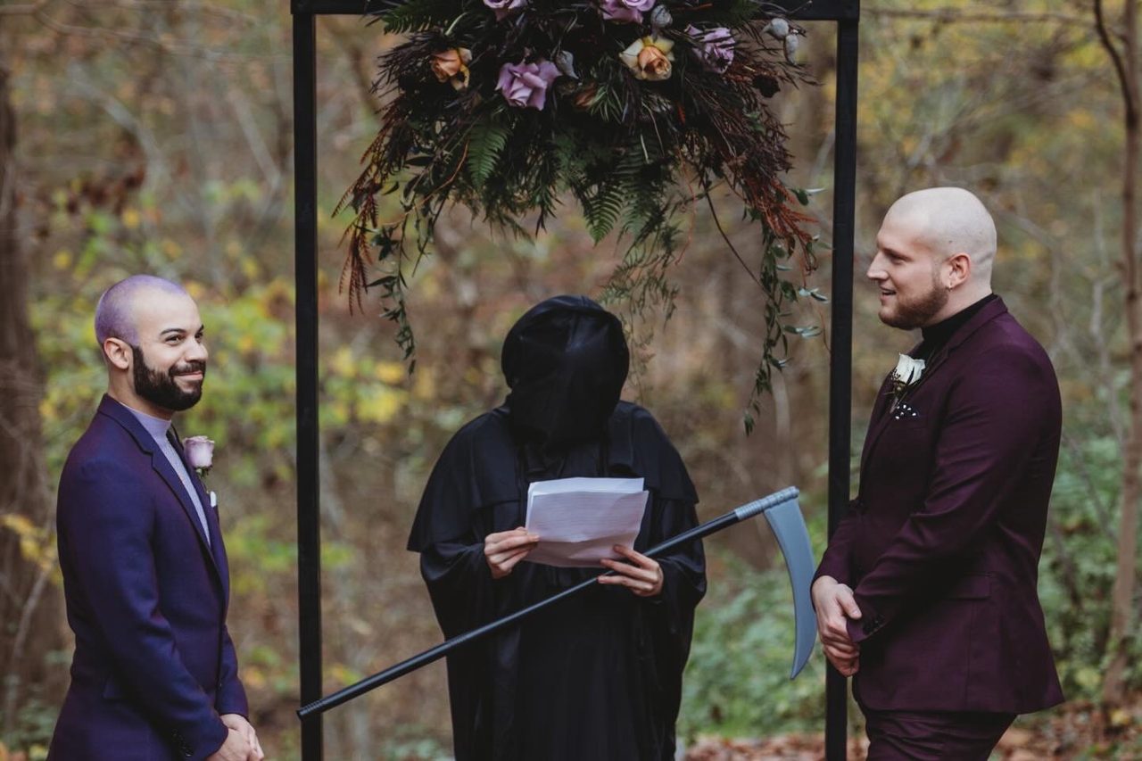 Xavi and Jared smiling at each other beneath a dark floral arch at their autumn wedding, while a friend officiates dressed as the Grim Reaper.